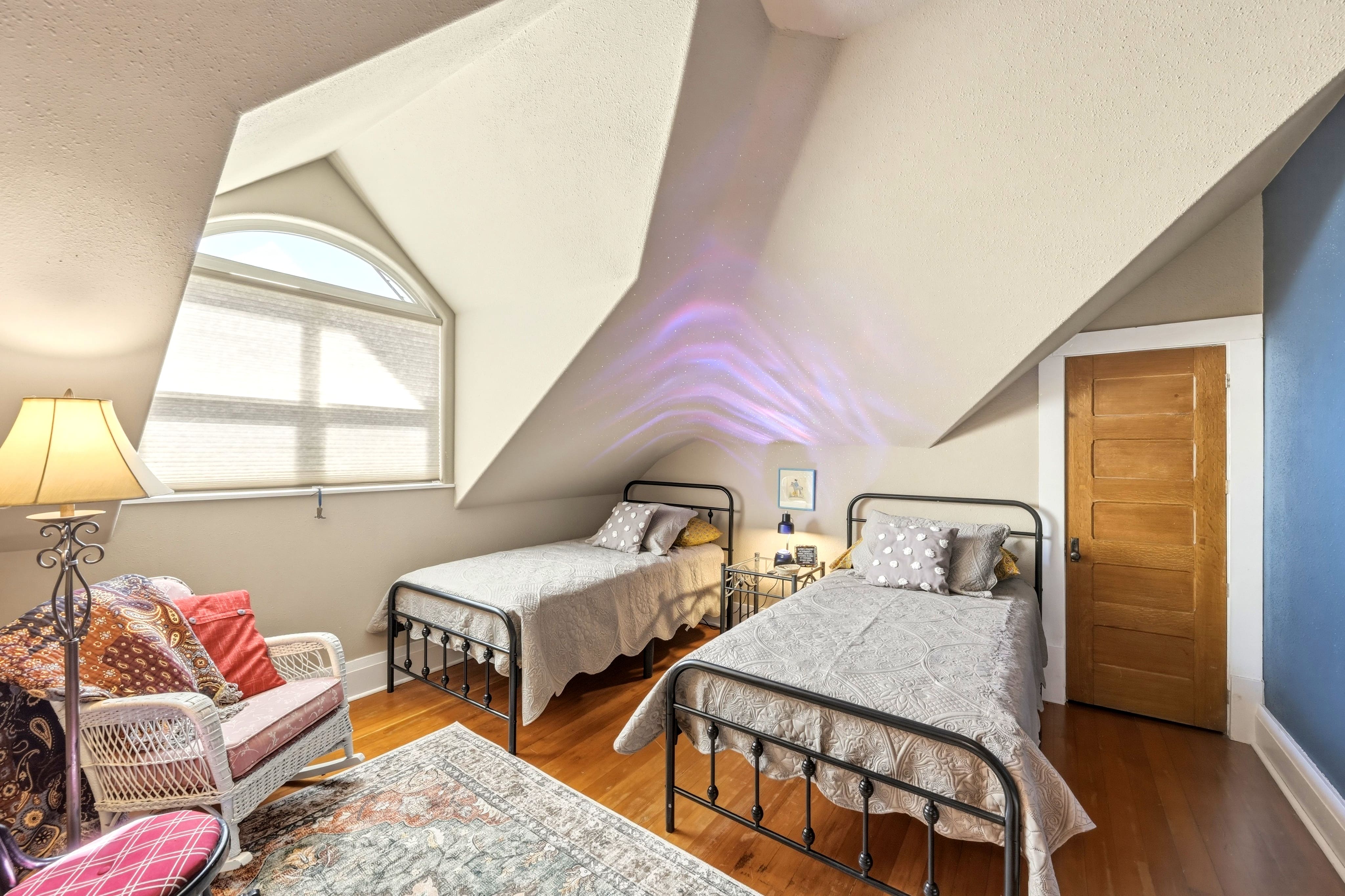 Bedroom with two twin beds, hardwood floor, sloping ceiling with arched window, and northern lights projected on ceiling.
